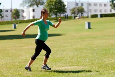Gravid kvinna springer i gräset, klädd i grönt linne och svarta tights. Hon verkar njuta av att lyfta gravid och vara aktiv utomhus.