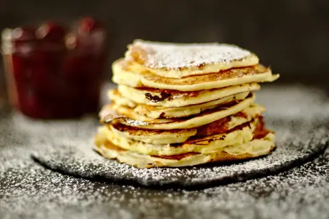 Ein Stapel fluffiger Quark Pfannkuchen, bestäubt mit Puderzucker, neben einem Glas Marmelade.