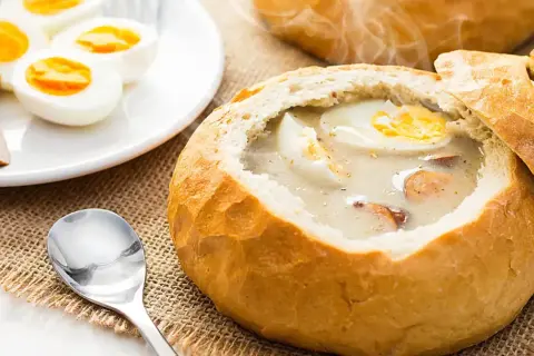 Żurek w chlebie z jajkiem i kiełbasą, obok talerz z jajkami i kiełbasą.