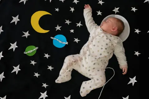 Śpiące dziecko w piżamce w kropki, otoczone gwiazdami i planetami, jakby leciało w kosmos. Ach śpij kochanie tekst pasuje do tej sceny.