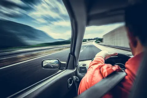 Kierowca w pomarańczowej kurtce jedzie autostradą. Widać rozmycie drogi i krajobrazu, co sugeruje dużą prędkość. Wymaga to ostrożności, a nawet szczególnej ostrożności.