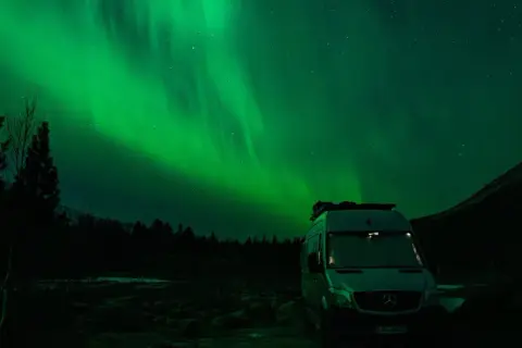 Ein Camper steht unter dem faszinierenden Licht der Aurora Borealis. Grüne Schleier tanzen am sternenklaren Himmel über dunklen Bäumen und einer Bergflanke.