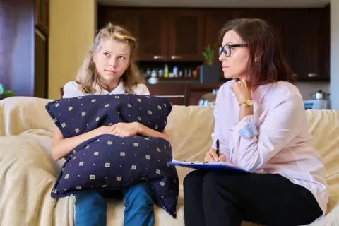 Dziewczynka z poduszką rozmawia z kobietą, która notuje. Może to być rozmowa o tym, ile zarabia kurator sądowy.