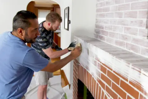 Two men are learning how to whitewash brick on a fireplace, applying white paint with brushes for a rustic look.