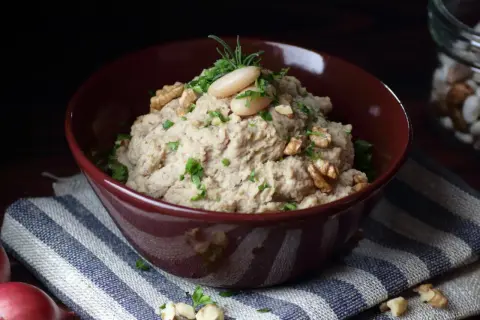 Aksamitna pasta z lubczyku, udekorowana fasolką, orzechami i ziołami, w bordowej misce na pasiastym obrusie.