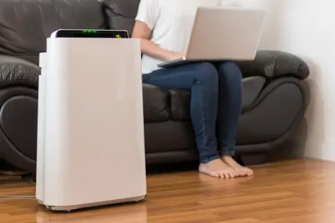 Oczyszczacz powietrza w salonie, obok osoby pracującej na laptopie. Dowiedz się, jak często wymieniać filtr w oczyszczaczu powietrza, by cieszyć się czystym powietrzem.