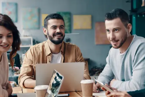 En grupp unga, leende människor diskuterar vid ett bord. En laptop och kaffekoppar antyder ett möte om utbildning, organisation och ledarskap.