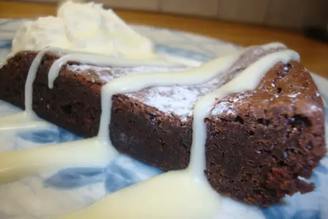 En bit mörk kladdkaka med pudersocker och vit chokladsås, serverad med vispad grädde.