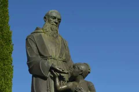 Escultura de fray Leopoldo con un niño, bajo un cielo azul. El fraile, con barba y hábito, extiende su mano hacia el pequeño.
