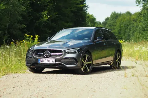 Szary Mercedes-Benz Klasy C All-Terrain na szutrowej drodze, otoczony zielenią lasu i polnymi kwiatami.