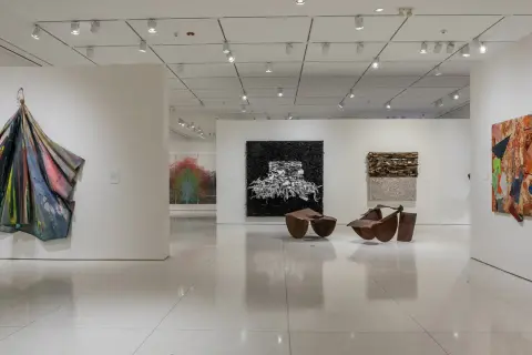 An art gallery features diverse works, including a colorful draped sculpture, a black and white textured piece, and a vibrant collage, reminiscent of the bold style of Pamela Joyner.