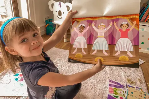 Uśmiechnięta dziewczynka pokazuje książeczkę z naklejkami baletnic. Idealny prezent na zajączka dla 3 latka, który uwielbia tańczyć.
