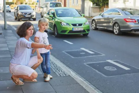 Mutter erklärt ihrer Tochter, dass sie auf dem Gehweg nicht in falsche Richtung fahren darf. Ein grünes Auto fährt auf der Straße.
