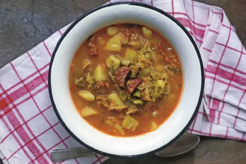 Gęsta zupa z kapusty włoskiej z ziemniakami i kiełbasą, podana w białej misce na kratkowanej serwetce.