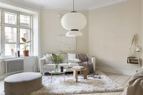 Ljust vardagsrum med en vit soffa, fluffig matta och en stor taklampa. Naturinredning med växter och trädetaljer skapar en lugn atmosfär.