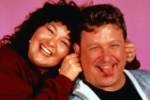 Roseanne und Dan, die Hauptfiguren der besetzung von Roseanne, lachen und Grimassen vor einem rosa Hintergrund.