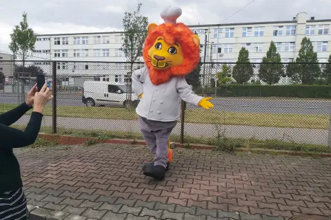 Uśmiechnięty strój lwa dla dziecka, w stroju kucharza, pozuje do zdjęcia na tle budynku.