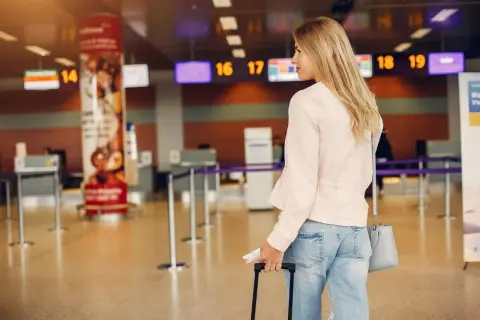 Odprawa na lotnisku w Katowicach: młoda kobieta z walizką i dokumentami czeka przy stanowisku odprawy, widoczne tablice z numerami lotów.