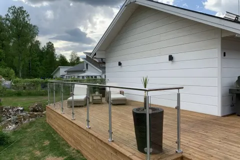 Träaltan med glasräcke och utemöbler. Huset är målat vitt, och det ser ut som att det nyligen har fått en ny omgång med olja på staketet.