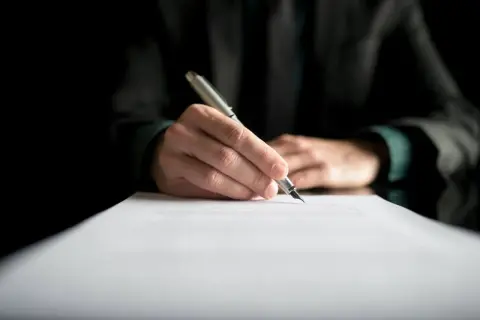 Dłoń pisze piórem na dokumencie. Może to być dziennik budowy, przerwa we wpisach lub ważny kontrakt.