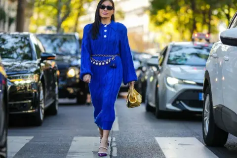 Elegancka kobieta w chabrowej sukience i fioletowych sandałkach na obcasie, z złotą torebką. Idealne buty do chabrowej sukienki to te, które podkreślą jej urok.