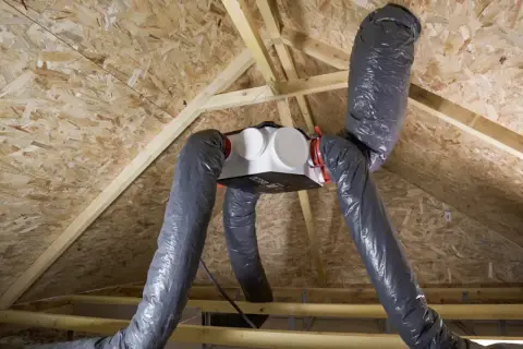 Installation d'une VMC dans un grenier. Le caisson blanc est suspendu, relié à des gaines grises flexibles.