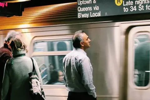 Personas esperando un tren en el metro de Nueva York con un letrero de destino.