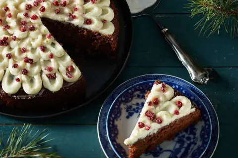 En mjuk pepparkaka med frostning och röda bär. En bit är upplagd på en blåvit tallrik, redo att avnjutas.