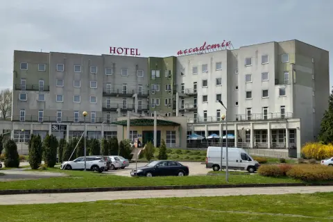 Hotel Accademia w Przemyślu, budynek z czerwonym napisem, parking z samochodami i zielony trawnik.