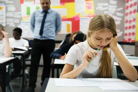 Młoda abiturientka zamyślona nad arkuszem egzaminacyjnym, podczas gdy nauczyciel obserwuje klasę.