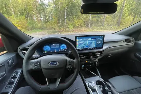 Wnętrze Ford Kuga z nowoczesnym ekranem multimedialnym i cyfrowymi zegarami. Kierownica z czerwonymi przeszyciami i przyciskami.