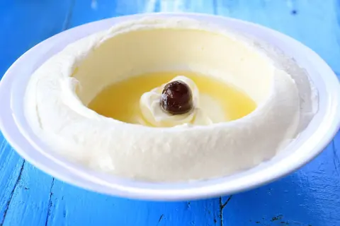 Un plato blanco con labneh que es un queso cremoso, con aceite de oliva y una aceituna negra en el centro, sobre una mesa azul.