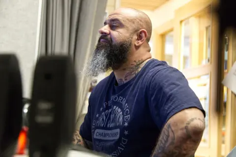 Man med skägg och tatueringar, klädd i en t-shirt med texten "Performance First Session Championship". Kanske en del av "kaka kartellen" som förbereder sig för något.