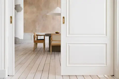 Puertas blancas y suelo de madera clara se combinan para crear un espacio luminoso. Al fondo, un comedor con sillas de madera y una mesa de hormigón.