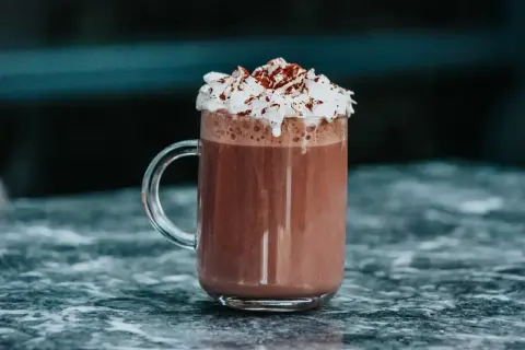 Un réconfortant chocolat chaud chantilly, garni de cacao, dans un mug en verre sur un marbre sombre.