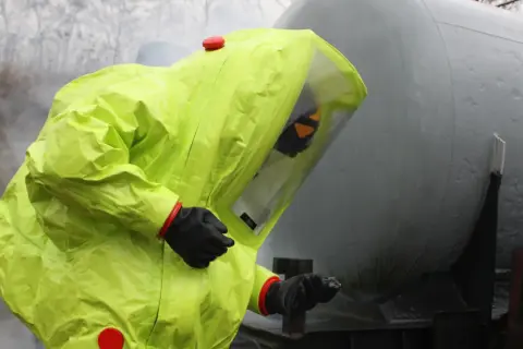 Person in a bright yellow hazmat suit with a clear visor, standing next to a large grey tank.