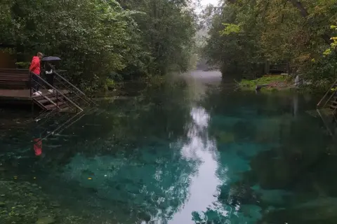 Turkusowe jezioro w Rosji, otoczone drzewami. Dwie osoby stoją na schodach prowadzących do wody, jedna z parasolem.