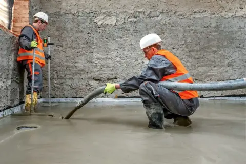 Dwóch budowlańców w kamizelkach odblaskowych wylewa pianobeton. Jeden z nich kieruje wąż, drugi kontroluje poziom.