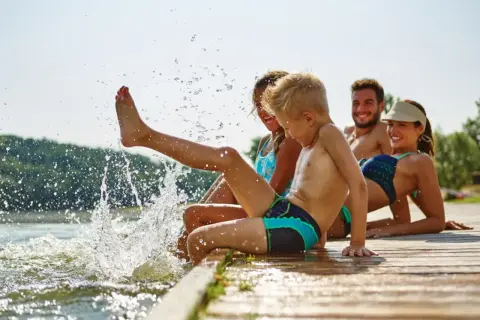 Familie genießt den Sommer am See. Kinder planschen im Wasser, Eltern entspannen sich. Guter uva schutz ist wichtig.