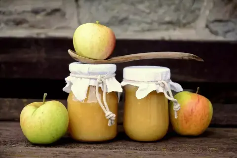 Deux pots de compote de pommes faits maison, décorés de tissus et de cordes, avec des pommes fraîches et une cuillère en bois.