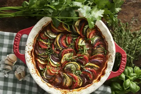 Vacker ratatouille i en röd ugnsform, med skivade grönsaker som tomat, zucchini och aubergine. Perfekt för ett ratatouille recept.