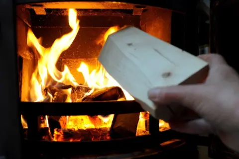 Holzscheit wird in einen brennenden Kamin gelegt. Vorsicht, keine Spanplatten verbrennen!