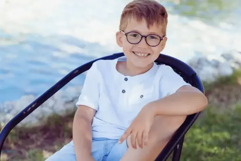 Uśmiechnięty chłopiec w okularach, 7 latek, siedzi na krześle przy wodzie.
