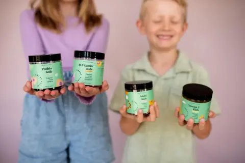 Två barn håller upp burkar med vitaminer. En flicka håller Probio Kids och D-Vitamin Kids, medan en pojke håller Multi Kids och Omega 3 Kids. Perfekt för vitamin barn!