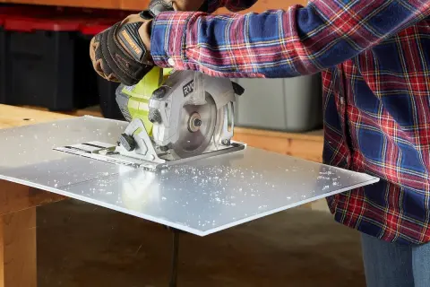 A person in a plaid shirt and work gloves uses a circular saw to cut a sheet of plexiglass, demonstrating how to cut plexiglass cleanly.