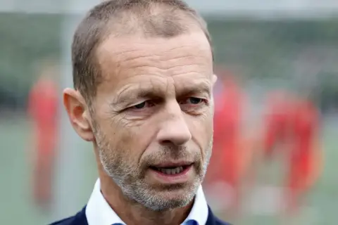 Aleksander Čeferin, prezes UEFA, w niebieskiej koszuli i granatowym swetrze, z rozmytym tłem piłkarzy.