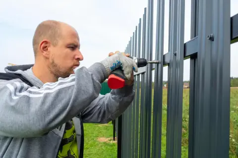 Mężczyzna w rękawiczkach przykręca wkrętarką element do nowego, szarego płotu.