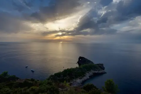 Atardecer en el mirador de Sa Foradada, con rayos de sol filtrándose entre nubes y dos barcos en el mar.