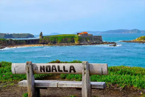 Banco de madera con #NOALLA grabado, frente al mar azul y un pueblo costero con edificios históricos.