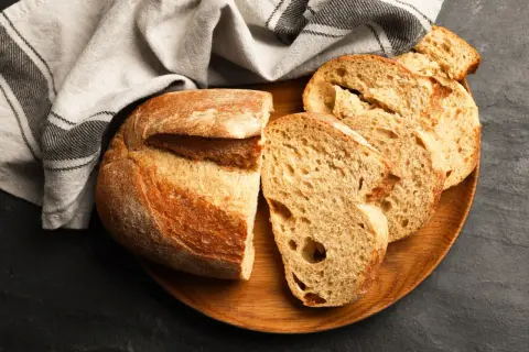 Krojony bochenek chleba i kromki na drewnianej desce, obok lnianej ściereczki.